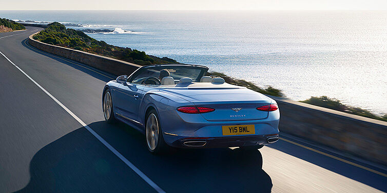 Bentley Tunbridge Wells New Bentley Continental GTC Azure convertible rear view in Light Windsor blue paint with 22 inch ten spoke wheels driving dynamically on an ocean road in the sunshine
